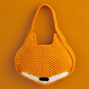 May include: A crochet handbag shaped like a fox head, crafted from vibrant orange yarn. The design features a single top handle, pointed ears, a white muzzle section at the bottom, and a small, solid black oval nose centred on the white area.
