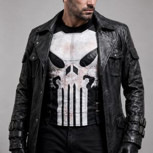 May include: Black leather jacket with a long design, featuring multiple pockets and buckle details. Underneath, a white and black graphic tee displays a skull design. The jacket is worn over a black shirt and black pants.