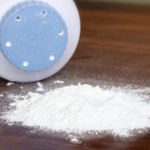 May include: A white baby powder container with a light blue lid and a pile of white powder on a wooden surface. The container has several small holes in the lid.