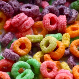 Può includere: Primo piano di cereali colorati a forma di anello in una ciotola. I cereali sono di vari colori, tra cui rosso, arancione, verde, giallo e viola. I cereali sono in un liquido bianco, probabilmente latte.