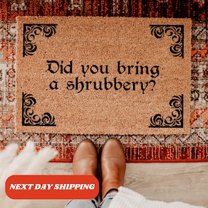 May include: A rectangular tan coir doormat featuring black decorative scrollwork in each corner and the black text "Did you bring a shrubbery?" centered on the mat. The mat has a coarse, natural fiber texture and a clean, simple design.
