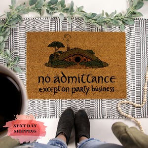 May include: A rectangular tan coir doormat featuring a black illustration of a round-door hillside dwelling with a chimney and trees. The text "no admittance except on party business" is printed in black in a stylized font across the center.