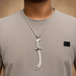 Puede incluir: Un collar de tono plateado con un colgante largo en forma de espada curva, con una superficie texturizada y una pequeña piedra preciosa redonda de color rojo oscuro engastada en el centro de la guarda. Cuelga de una cadena fina trenzada.