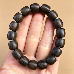 Puede incluir: Una pulsera de cuentas de madera marrón oscuro. La pulsera está compuesta por cuentas en forma de barril ensartadas. Las cuentas tienen un acabado liso y pulido y un color rico y oscuro. La pulsera se sostiene en una mano sobre un fondo neutro.