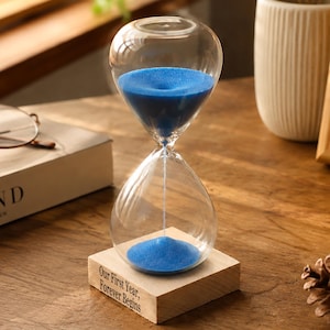 May include: A clear glass hourglass filled with vibrant blue sand, mounted on a square light-coloured wooden base. The base features the engraved text, "Our First Year, Forever Begins," positioned on the front side of the wooden block.