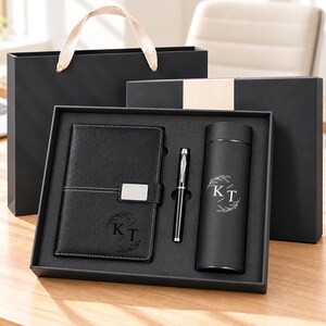 May include: A black gift set containing a notebook with a silver metal clasp, a black ballpoint pen with silver accents, and a black cylindrical vacuum flask. Both the notebook and the flask feature the engraved letters KT surrounded by a leaf wreath.