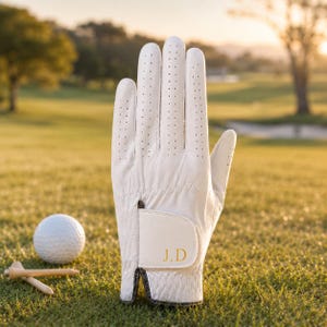 Peut inclure: Un gant de golf blanc avec de petites perforations et les initiales "J.D" en or. Une balle de golf et deux tees sont sur l'herbe verte. Le gant est sur l'herbe, avec un arrière-plan flou d'un terrain de golf.