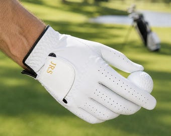 May include: A white golf glove with black trim and the initials "JRS" in gold lettering. The glove is holding a white golf ball. The background shows a golf course with green grass and a lake.