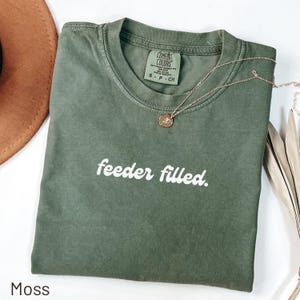 Puede incluir: Una camiseta verde musgo con la frase "feeder filled" en escritura blanca. Un collar dorado con un colgante descansa sobre la camiseta. La camiseta está doblada sobre una superficie blanca, con un sombrero marrón visible en el fondo.