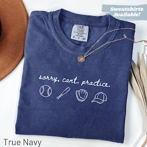 May include: A true navy blue t-shirt featuring the white text "sorry, cant. practice." above white line art illustrations of a baseball, a baseball bat, a baseball glove, and a baseball cap. The shirt has a Comfort Colors brand label.