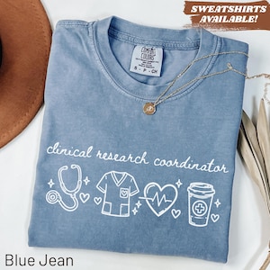 May include: Blue jean colored t-shirt with the words "clinical research coordinator" and medical-themed graphics. The shirt has a crew neck and short sleeves. A gold necklace with a pendant is on the shirt. The text "SWEATSHIRTS AVAILABLE!" is in the top right corner.