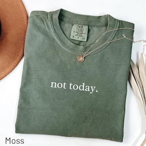 Könnte beinhalten: Ein moosgrünes T-Shirt mit dem weißen Aufdruck "not today". Eine goldene Halskette mit Anhänger liegt auf dem Shirt. Das Shirt ist gefaltet und auf einer weißen Oberfläche platziert.