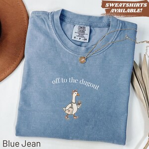 May include: Blue jean colored t-shirt with the text "off to the dugout". The shirt features an embroidered cartoon goose wearing a pink bow, carrying a coffee cup and a tote bag. A gold necklace is draped across the shirt.