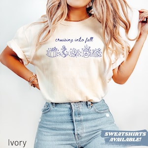 May include: An ivory short-sleeved t-shirt featuring the dark blue text "cruising into fall" above a line of icons including a pumpkin, an anchor, a maple leaf, a steaming mug, and a wave. The shirt is paired with light blue denim jeans.