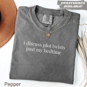 Könnte beinhalten: Ein anthrazitfarbenes Kurzarm-T-Shirt mit dem weißen Text "i discuss plot twists past my bedtime" auf der Vorderseite. Das T-Shirt hat einen Rundhalsausschnitt und ein Markenetikett von Comfort Colors im Kragenbereich. Eine goldene Kette liegt darauf.