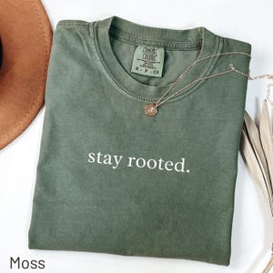 May include: A moss green t-shirt with the words "stay rooted" printed in white. A gold necklace with a circular pendant lies on the shirt. The shirt is folded, displayed with a brown hat and decorative leaves.