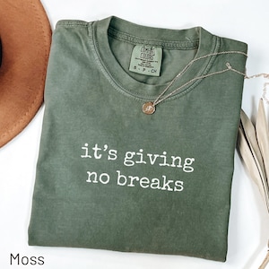 以下が含まれることがあります： 「it's giving no breaks」というフレーズが白でプリントされたモスグリーンのTシャツ。ペンダント付きのゴールドネックレスがシャツの上にあります。茶色の帽子が左上にあります。シャツは白い表面に折りたたまれています。