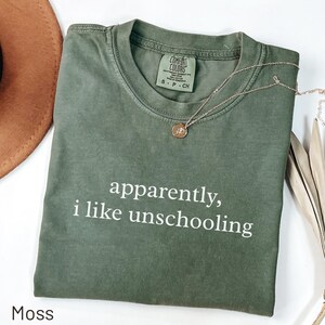 May include: A moss green short-sleeve t-shirt with a crew neckline and the white text "apparently, i like unschooling" printed on the front. The shirt features a Comfort Colors brand label inside the collar and a thin gold-toned necklace.