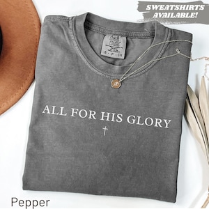 Puede incluir: Una camiseta gris Comfort Colors con el texto "ALL FOR HIS GLORY" y una cruz. Un collar de oro con un colgante descansa sobre la camisa. Las palabras "SWEATSHIRTS AVAILABLE!" están en la esquina superior derecha.
