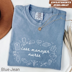 May include: A blue jean colored t-shirt with a white graphic print featuring the text case manager nurse surrounded by medical icons including a stethoscope, heart, face mask, clipboard, IV bag, thermometer, and gloves, with a gold necklace.