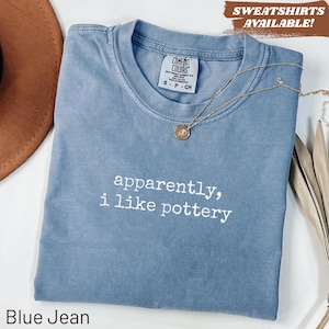 May include: A blue jean colored t-shirt with a crew neckline and the white text "apparently, i like pottery" printed on the front. A gold-toned necklace with a small circular pendant rests on the collar area of the garment.