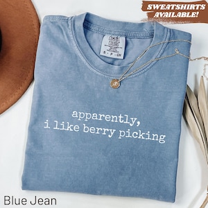 May include: A blue jean colored t-shirt with a crew neckline and a Comfort Colors brand label. The shirt features the white text "apparently, i like berry picking" printed on the front. A gold-toned necklace with a small circular pendant rests on the shirt.