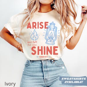 May include: Ivory-colored t-shirt with the words "ARISE SHINE" in red, and a blue illustration of a lantern and flames. The text "your light has come" is below the flame graphic. The shirt also has the text "ISAIAH 60:1".