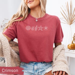 May include: Crimson-colored t-shirt with a crew neck and short sleeves. The shirt features a white line drawing of seashells, a starfish, and a compass rose. The word "Crimson" is displayed in a red box at the bottom of the image.