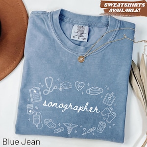 Puede incluir: Camiseta azul vaquero con la palabra "sonographer" en escritura blanca, rodeada de ilustraciones médicas. La camiseta tiene cuello redondo y mangas cortas. El texto "SWEATSHIRTS AVAILABLE!" está en la esquina superior derecha.