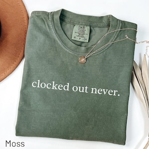 May include: A moss green t-shirt with the phrase "clocked out never" in white. The shirt has a crew neck and short sleeves. A gold necklace with a pendant rests on the shirt. The shirt is folded on a white surface.