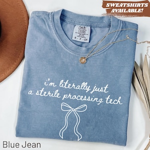 Op de afbeelding: Een blauw spijkerkleurig t-shirt met een ronde hals en een Comfort Colors label. Het shirt heeft witte tekst met de tekst "i'm literally just a sterile processing tech" boven een witte lijntekening van een strik. Een gouden ketting ligt op het shirt.