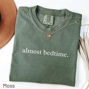 May include: A moss green t-shirt with the phrase "almost bedtime" printed in white. A gold necklace with a pendant rests on the shirt. The shirt is folded and displayed with a brown hat and decorative leaves.