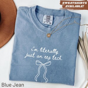 May include: A blue jean coloured t-shirt featuring the white text "i'm literally just an eeg tech" above a simple white line drawing of a bow. The shirt has a crew neckline and a Comfort Colors brand label visible at the inner collar.