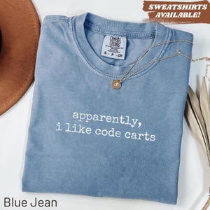 May include: A blue jean colored t-shirt with a crew neckline and a Comfort Colors brand label. The shirt features the white text "apparently, i like code carts" printed on the front. A gold-toned necklace with a small circular pendant rests on the fabric.