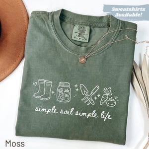 May include: A moss green t-shirt featuring a white graphic of gardening tools, wellington boots, and vegetables. The text "simple soil simple life" is printed below the graphic. A gold necklace is draped across the shirt. The shirt is labeled "Comfort Colors".