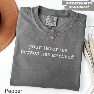 May include: A charcoal gray short-sleeve t-shirt with a crew neckline and the white text "your favorite person has arrived" printed on the chest. The shirt features a Comfort Colors brand label at the inner neck and a delicate gold-tone chain necklace.