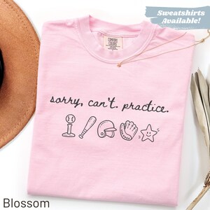 Op de afbeelding: Een lichtroze T-shirt met korte mouwen en de zwarte tekst "sorry, can't. practice." boven een rij zwarte lijntekeningen van een honkbal op een tee, een honkbalknuppel, een helm, een honkbalhandschoen en een lachend sterretje.