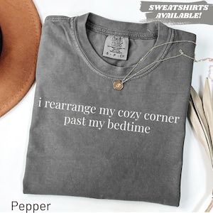 May include: A charcoal gray short-sleeve t-shirt with the white text "i rearrange my cozy corner past my bedtime" printed on the front. The shirt features a crew neckline and a Comfort Colors brand label inside the collar. A gold necklace is visible.