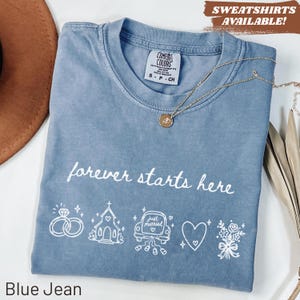 Puede incluir: Camiseta azul vaquero con la frase "forever starts here" y gráficos temáticos de boda. Un collar de oro con un colgante descansa sobre la camisa. La camisa tiene una etiqueta Comfort Colors.