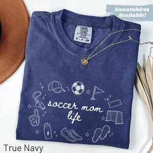 Può includere: T-shirt blu navy con le parole "soccer mom life" e grafica correlata in bianco. La maglietta ha girocollo e maniche corte. Il testo "Sweatshirts Available!" si trova nell'angolo in alto a destra.