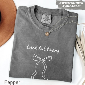 Puede incluir: Una camiseta gris con la frase "tired but trying" y un lazo blanco. Un collar dorado con un colgante descansa sobre la camiseta. La camiseta está etiquetada como "Pepper" en la parte inferior.