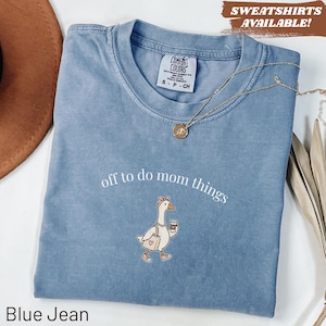 May include: Blue jean colored t-shirt with the phrase "off to do mom things" and a cartoon goose carrying a coffee cup. The shirt has a crew neck and short sleeves. A gold necklace is draped across the shirt. The text "SWEATSHIRTS AVAILABLE!" is in the top right corner.