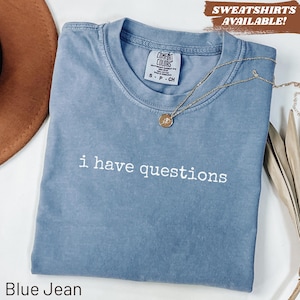 May include: A blue jean Comfort Colors t-shirt with the phrase "i have questions" in white. A gold necklace with a pendant rests on the shirt. The shirt is displayed on a white surface.