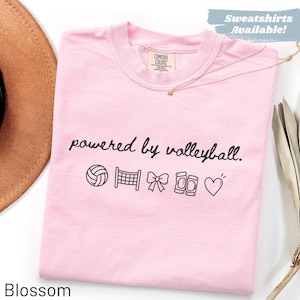 May include: A light pink t-shirt with the phrase "powered by volleyball" and volleyball-themed graphics. The shirt has a crew neck and a tag that says "Comfort Colors". The word "Blossom" is at the bottom.