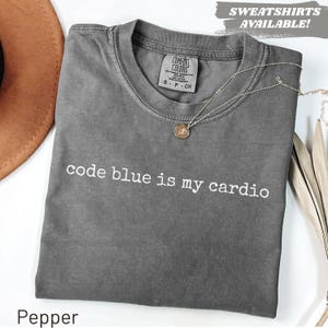 May include: A gray t-shirt with the text "code blue is my cardio" in white. The shirt has a crew neck and short sleeves. A gold necklace with a pendant is draped across the shirt. The word "Pepper" is at the bottom of the image.