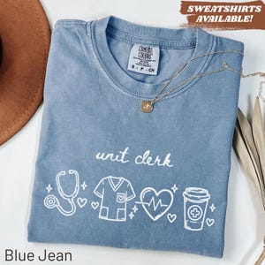 May include: A blue jean coloured t-shirt featuring the words "unit clerk" and medical-themed illustrations. The illustrations include a stethoscope, scrubs, a heart with a heartbeat line, and a coffee cup. A gold necklace is draped across the shirt.