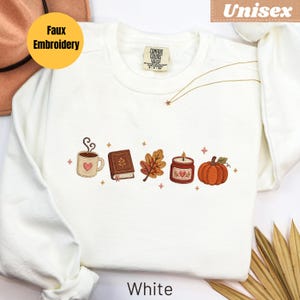 Könnte beinhalten: Ein weißes Langarm-Sweatshirt mit einer Faux-Stickerei-Grafik, die eine dampfende Kaffeetasse mit Herz, ein geschlossenes Buch, ein Herbstblatt, eine brennende Kerze und einen orangefarbenen Kürbis zeigt, akzentuiert durch kleine Sternformen.