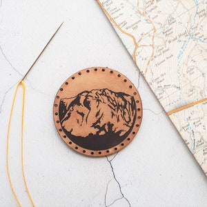 May include: A circular, tan leather patch featuring a mountain range design. The patch is hand-stitched around the edge with small, evenly spaced holes. A needle with yellow thread and a vintage map are also in the image.