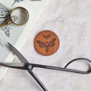 Puede incluir: Un círculo de cuero marrón con un diseño de polilla en blanco y negro. La polilla tiene una envergadura de aproximadamente 5 cm. El círculo está rodeado por una fila de pequeños agujeros. También hay un par de tijeras de metal negro en la imagen.