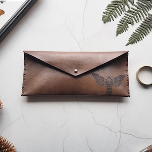 May include: A brown leather envelope clutch with a silver button closure. The clutch has a black moth design embossed on the front.